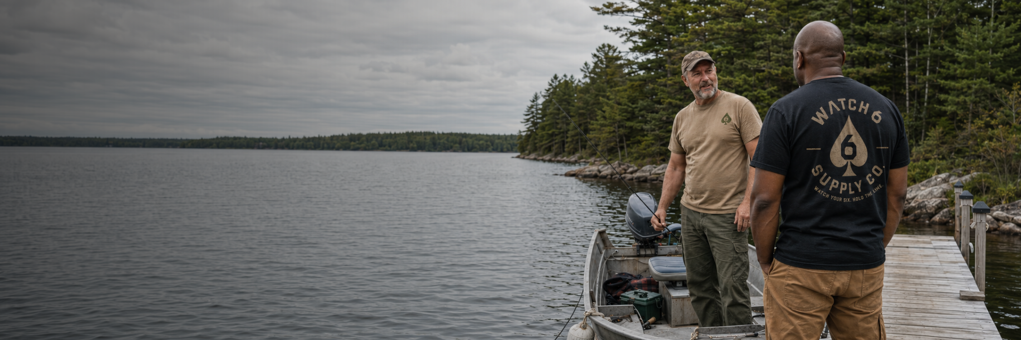 Two veterans wearing Watch 6 shirts beside a fishing boat on a wooded lake.